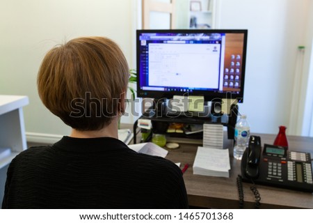 Person works on computer at office desk. A close up view of a lady worker sat a computer desk concentrating on PC screen. Back of head visible at a busy desk. Workplace employee carries out IT duties.