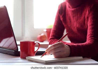 Person Writes In A Notebook Next To A Red Mug And A Laptop, Sitting In Front Of The Window / Cozy Working Atmosphere In Red Tones