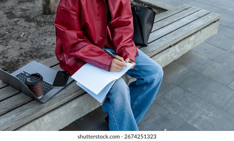 A person writes in a notebook while sitting on a park bench, with a laptop, phone, and coffee cup nearby.