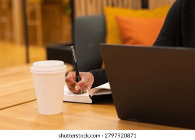 A person writes notes while working on a laptop, with a coffee cup placed on a wooden table. The warm, inviting setting suggests focus and productivity in a cozy workspace. This image conveys themes of diligence, creativity, and modern working environments, suitable for illustrating various professional or educational contexts.