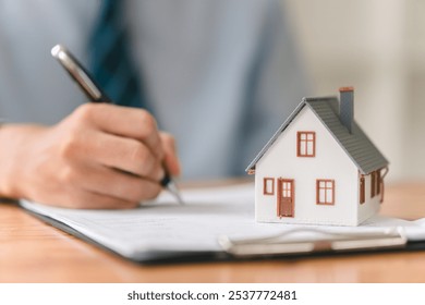 A person writes on a document next to a miniature house, symbolizing real estate transactions or home ownership activities.
