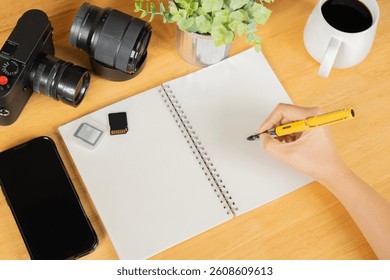 A person writing in a notebook with camera, memory cards, phone and lens on wooden table. concept of simple creative work space