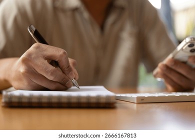 person writing notes in a notebook while holding a smartphone, concept for multitasking, productivity, planning, business organization, and creative work process.