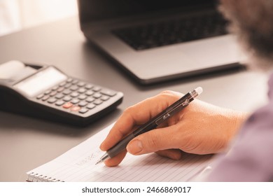 A person writing notes on a notepad with a pen, accompanied by a calculator and laptop on a home office desk. The setup highlights financial planning and modern workspace essentials. - Powered by Shutterstock