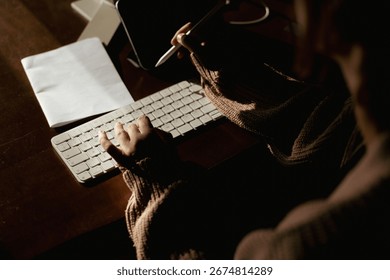 Person writing notes while typing on a keyboard in a cozy indoor setting during evening hours