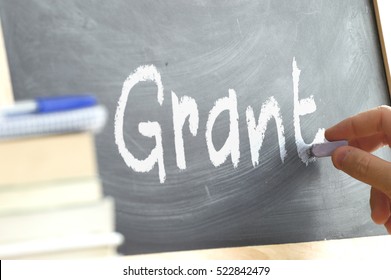 A Person Writing The Word Grant On A Blackboard. Some School Materials And Copy Space.