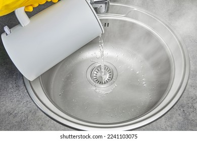 A person in yellow rubber gloves pours hot water with steam from an electric kettle into a metal sink. Cleaning and disinfection of sanitary ware in the kitchen - Shutterstock ID 2241103075