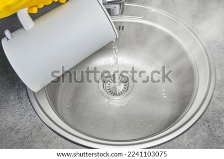 A person in yellow rubber gloves pours hot water with steam from an electric kettle into a metal sink. Cleaning and disinfection of sanitary ware in the kitchen
