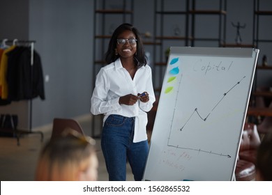 Personable African Woman In Eyeglasses, Wearing White Shirt And Jeans, Using Diagrams, Motivated African American Speaker Coach Present Business Plan On Whiteboard For Workers In Modern Office