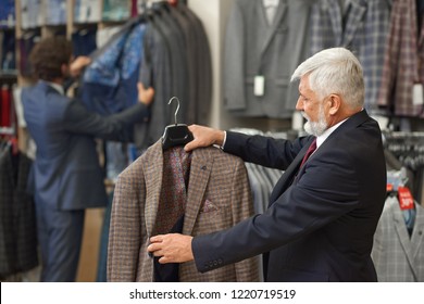 Personable Older Man With Grey Hair Holding Item And Looking At It. Customer Thinking About Buying Checkerd Brown Jacket. On Background Young Guy Choosing Clothes. Clients On Shopping In Boutique.