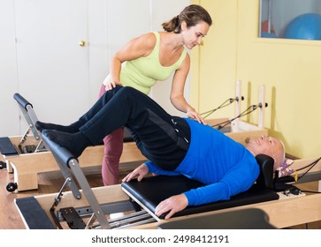 Personal female trainer controlling movements of senior man doing pilates on reformer in fitness studio. Healthy active lifestyle - Powered by Shutterstock