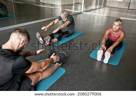 Personal fitness instructor and couple of elderly man and woman stretching on gym mats to relax the tension from the workout. Cooldown exercises for active healthy couple.