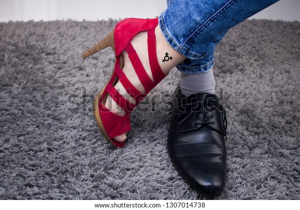 Personal has a lady`s boot on one foot, and on\
the other a male boot.The person has a third-gender tattoo.Man or\
woman? Person has woman´s red sandal and men´s black formal\
shoe.Gender conflict.