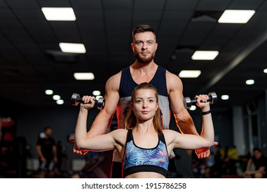 Personal Male Trainer Helps Woman Client With Physical Exercises In Gym. Dark Gym Background. Closeup. Front View.