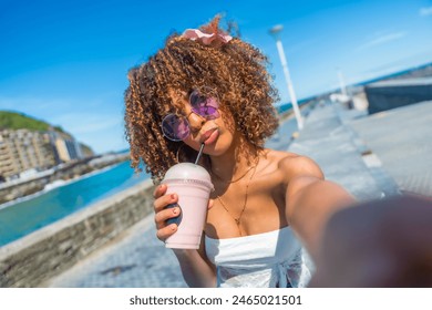 Personal perspective of a cool latin young woman with curly hair and sunglasses drinking milkshake taking a selfie on the promenade - Powered by Shutterstock