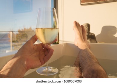 Personal Perspective Man Relaxing In Bath Tub With Glass Of White Wine In Hand By Sea View Window.