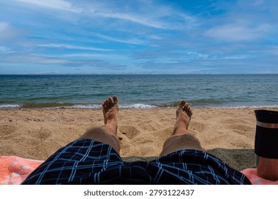 A personal perspective view of a man's sandy feet and legs in plaid swim trunks, sunbathing on a towel at the beach with a view of the calm blue sea, horizon, and sky.