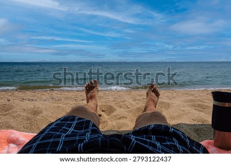 A personal perspective view of a man's sandy feet and legs in plaid swim trunks, sunbathing on a towel at the beach with a view of the calm blue sea, horizon, and sky.