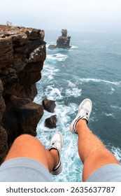 A personal point of view shot capturing legs and white sneakers hanging over the steep edge of a cliff at Cabo Carvoeiro, overlooking the ocean below