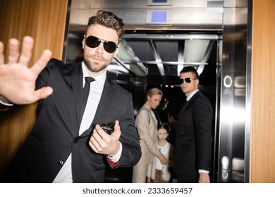 personal safety, handsome bodyguard in sunglasses and suit showing stop, no gesture to camera, protecting private space of famous clients in elevator of luxury hotel, blurred mother and daughter - Shutterstock ID 2333659457