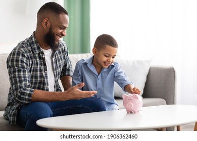 Personal Savings. African Father And Son Putting Coin In Piggybank Sitting On Sofa At Home, Dad Teaching His Kid Financial Literacy, Talking About Money And Budget Planning. Selective Focus