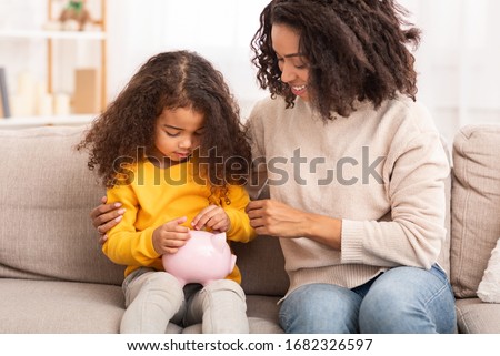 Personal Savings. Little African Girl Putting Money To Piggybank Sitting With Mother On Couch Indoor