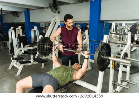 Personal trainer assisting man doing bench press exercise in gym. Focusing on fitness, strength training, and workout routine