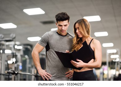 Personal Trainer And Client Looking At His Progress At The Gym. Athletic Man And Woman Wearing Sportswear.