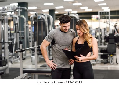 Personal Trainer And Client Looking At His Progress At The Gym. Athletic Man And Woman Wearing Sportswear.