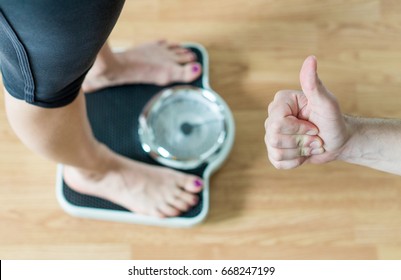 Personal Trainer Or Coach Giving Thumbs Up To A Person Standing On Scale. Success And Great Achievement In Weight Loss. Encouragement And Motivation. Happy And Satisfied With The Results.