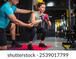 Personal trainer is helping a woman to keep her back straight while doing squats with a medicine ball in a gym