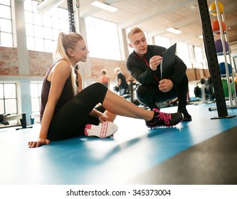 Personal trainer helping young woman on her work out routines in gym. Female sitting on floor with her personal trainer showing fitness report on a clipboard. - Powered by Shutterstock