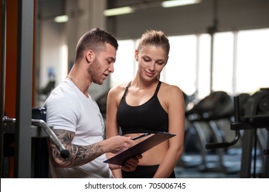 Personal Trainer And His Female Client Standing Together And Discussing Nutrition Or Training Plan On Clipboard. Cute Blonde Woman And Coach In Fitness Center. Healthy Lifestyle, Fitness Concept.