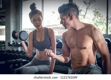 Personal trainer man teach Asian women`s to pull up dumbbell weights for exercise at indoor sport gym, bodybuilding and gym equipment concept.