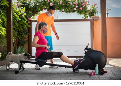 Personal Trainer With Protection Mask And Senior Woman Doing Rowing Following The Teacher's Instructions. Exercises At Home In Times Of Pandemic. Home Gym. Family Exercise