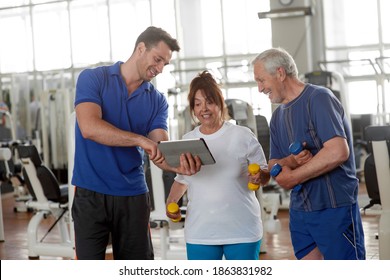 Personal trainer showing exercise to senior people in gym. Personal trainer using digital tablet while talking to senior couple. People, sport, technology, healthy lifestyle.