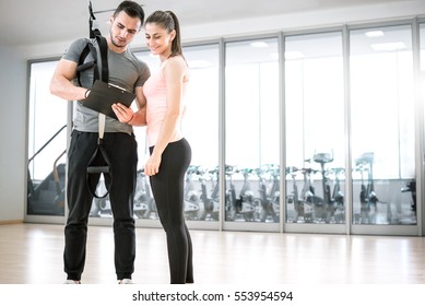 Personal Trainer Showing Result Of Training Plan To His Female Young Client With Suspension Rope Over Shoulder.