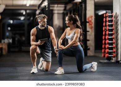 Personal trainer is showing a sportswoman how to do lunges correctly while kneeling in a gym.