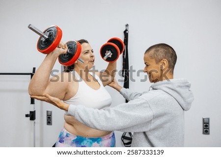 Personal trainer supporting client lifting dumbbells in gym for back and shoulder workout