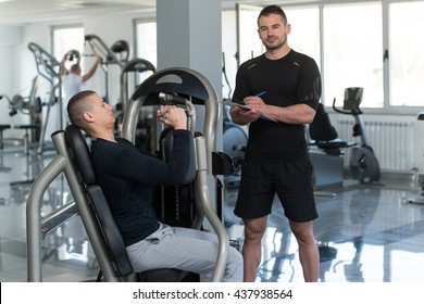 Personal Trainer Takes Notes While Young Man Exercise Chest On Machine In The Gym