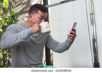 A personal trainer wearing a gray sweater read his messages on the phone while seated on the squat rack. Drinking coffee from a mug. At a home gym.