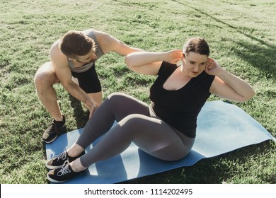 Personal Trainer Working With His Client Outdoors. Overweight Woman Doing Situps On Mat With Assistance Of Her Fitness Instructor Support. Sport, Training, Weight Loss, Teamwork, Lifestyle Concept.