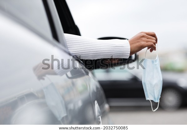 Personal transport, new normal, social distance,\
buying car, goodbye covid-19 pandemic. Hand of young african\
american female holds protective mask in open auto window, cropped,\
copy space, close up