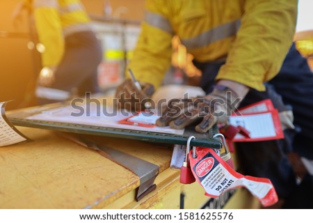 Personnel red danger locks attached with danger tags are locking on safety isolation permit lock box with defocused worker writing name, sign on prior locking on work permit safety control box