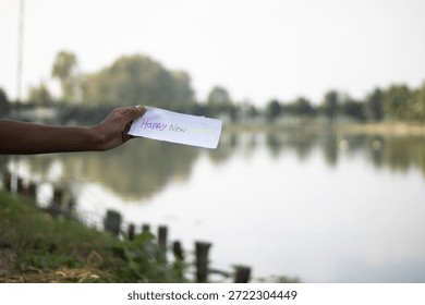 A person's arm extends from the left side holding a white piece of paper with blurry writing over a calm lake reflecting trees and a bright sky with a grassy bank and wooden posts