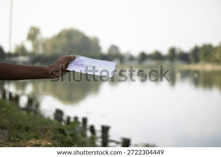 A person's arm extends from the left side holding a white piece of paper with blurry writing over a calm lake reflecting trees and a bright sky with a grassy bank and wooden posts
