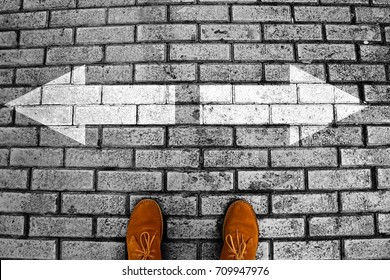 Person's Feet In Suede Shoes Is Standing At Tile Green Pavement Crossroad With White Arrows Print Pointing In Two Different Directions. Two Ways To Choose Making Decision Which Way To Go. Top View