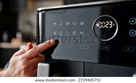 A person's finger is shown pressing a button on the digital interface of a sleek, black air fryer, selecting a cooking preset