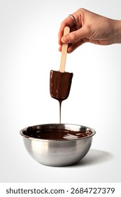 A person's hand, adorned with a ring, dips a wooden-stick popsicle into a stainless steel bowl filled with rich, melted chocolate.