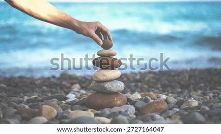 A person's hand carefully balances a stack of smooth stones on a rocky beach, with the ocean waves gently lapping in the background, creating a serene and peaceful atmosphere with shades of blue, brow